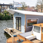 Exterior side close-up of a floating shepherd’s hut with waterside deck, large windows and floating platform.