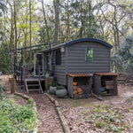 Side exterior view of a compact shepherd’s hut with woodland pathway, private terrace and rustic outdoor storage.