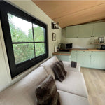 Open-plan kitchen and lounge inside The Elmstead 2 Person Shepherd’s Hut with large window, sofa and sage green cabinetry