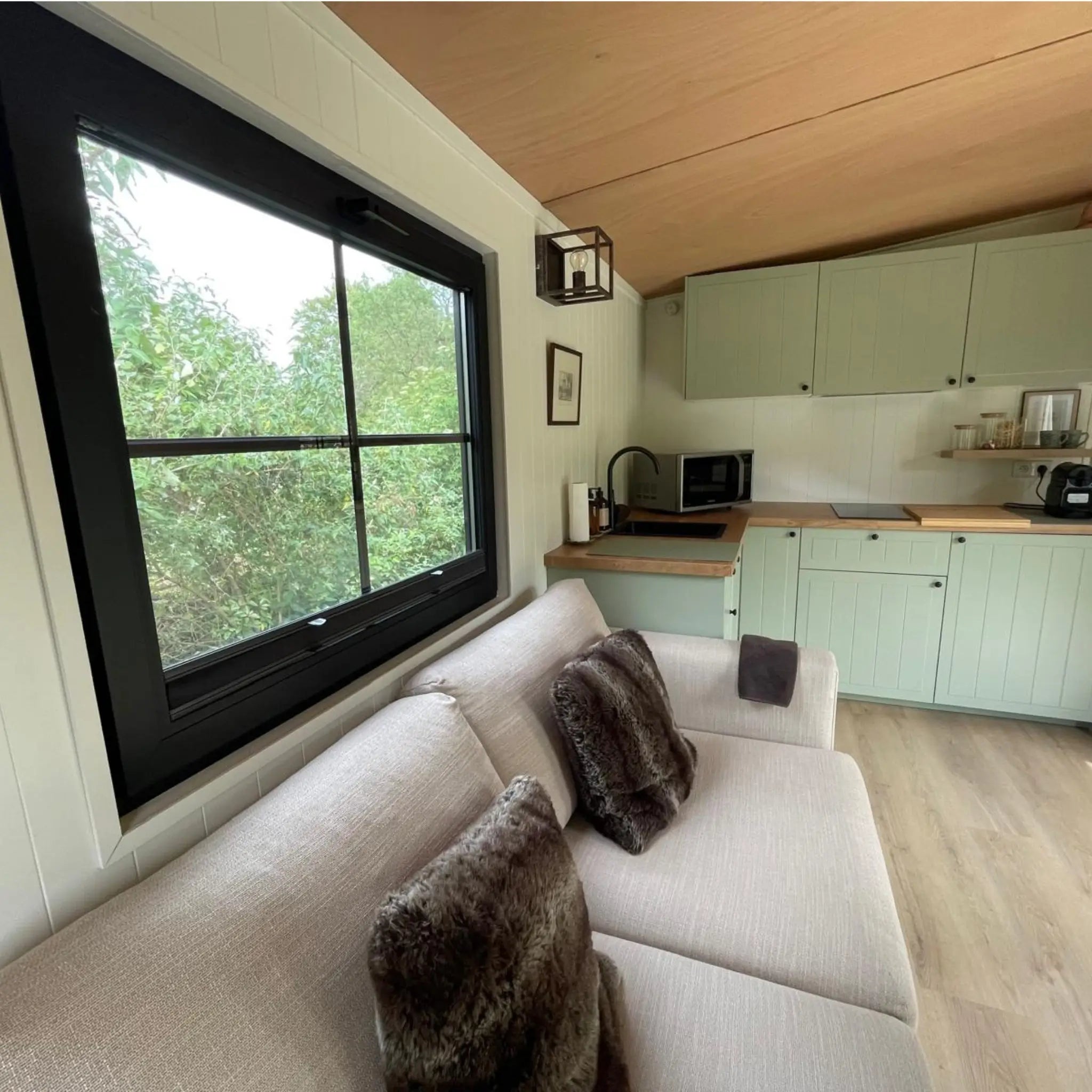 Open-plan kitchen and lounge inside The Elmstead 2 Person Shepherd’s Hut with large window, sofa and sage green cabinetry