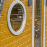 Close-up of circular window on wooden Grotto 3 person glamping pod showing natural timber exterior and modern design