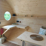The Grotto luxury 3 person glamping pod interior with timber finish, dining area, wall-mounted table and circular window.