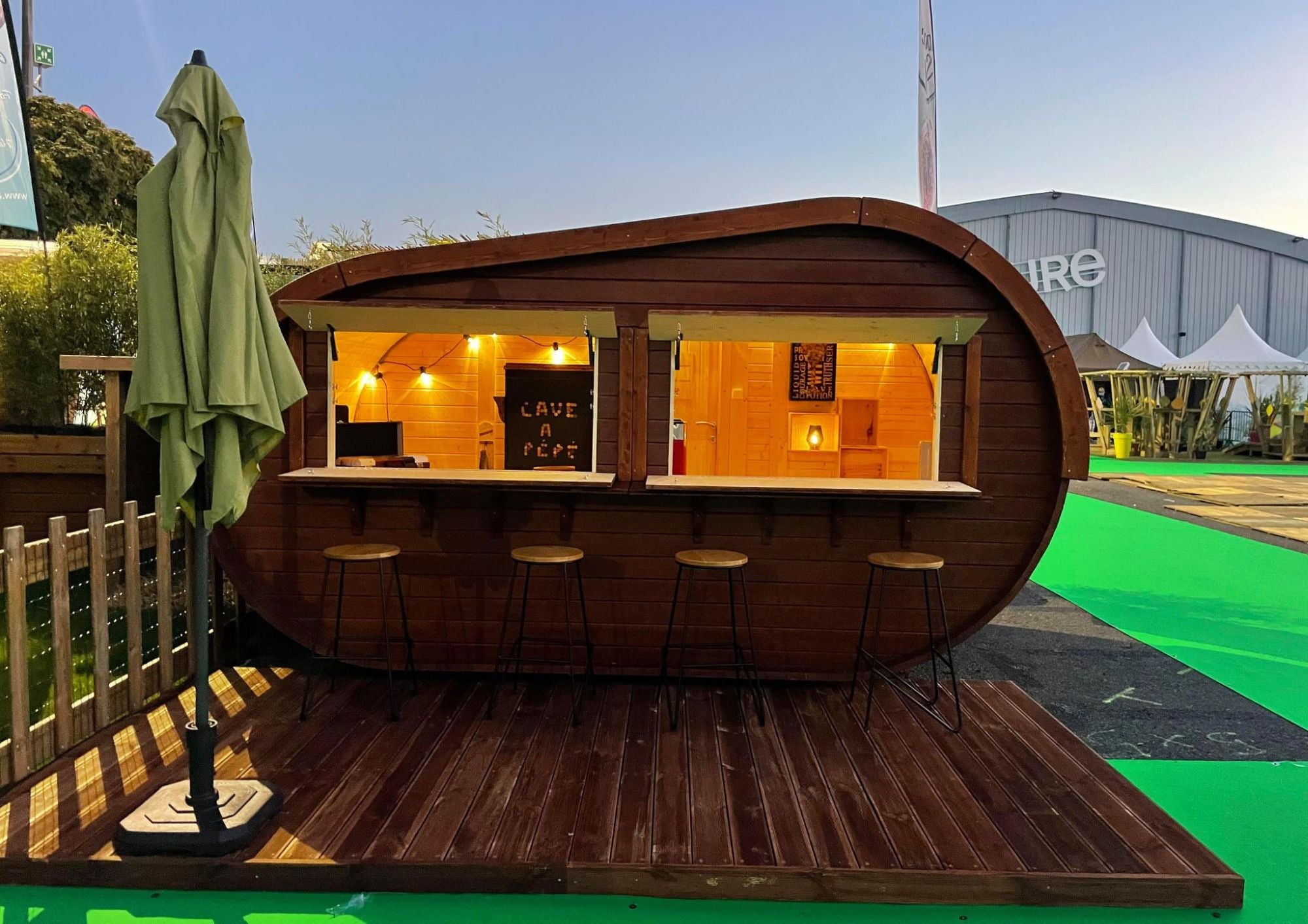 Wooden barrel-shaped bar with interior lights, outdoor furniture, and a green umbrella on a wooden deck.