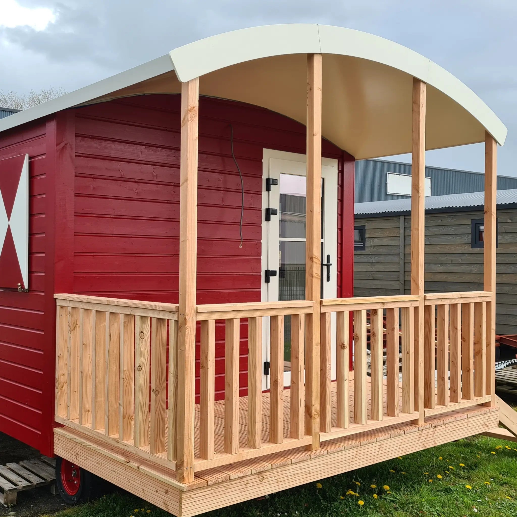 Front exterior view of The Hawthorne luxury shepherds hut with covered timber porch, curved roof and red cladding.