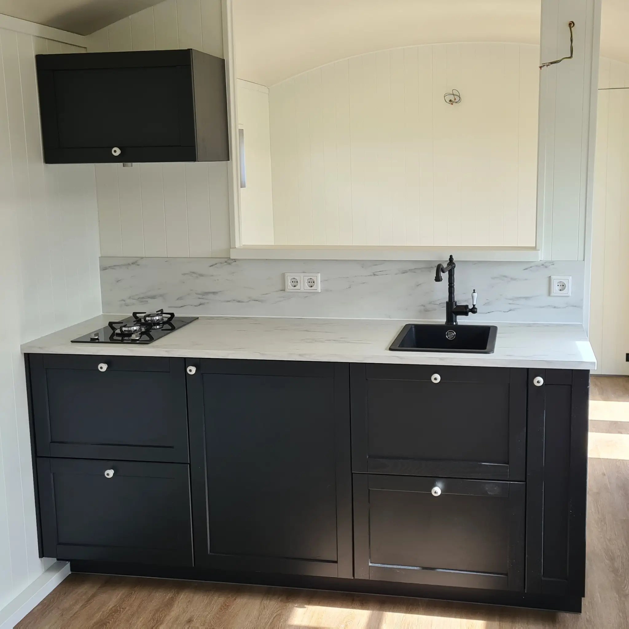 Compact kitchenette close-up in The Hawthorne shepherd hut featuring black cabinetry, marble-effect worktops and integrated gas hob.