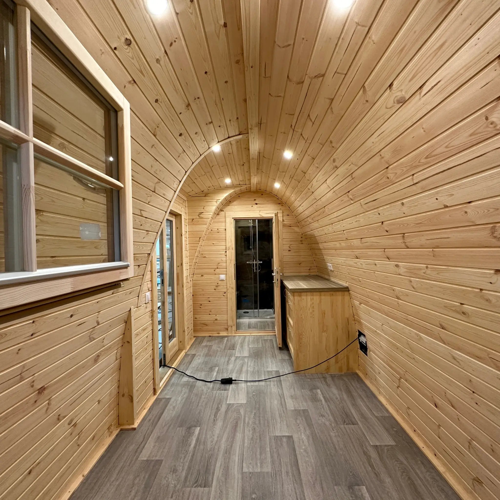 Interior view of The Hearthwood Pod showing open-plan living space with curved timber ceiling, wood-clad walls, and vinyl flooring