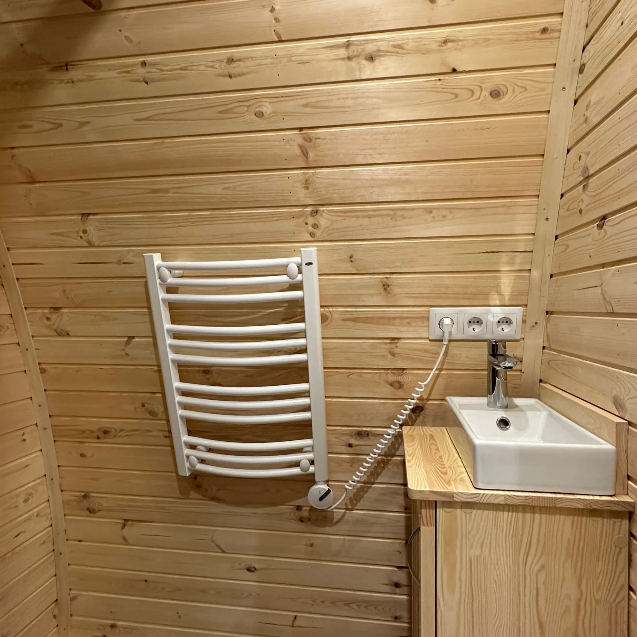 Compact glamping pod bathroom with electric towel radiator, timber wall cladding, and modern sink unit in The Hearthwood Pod
