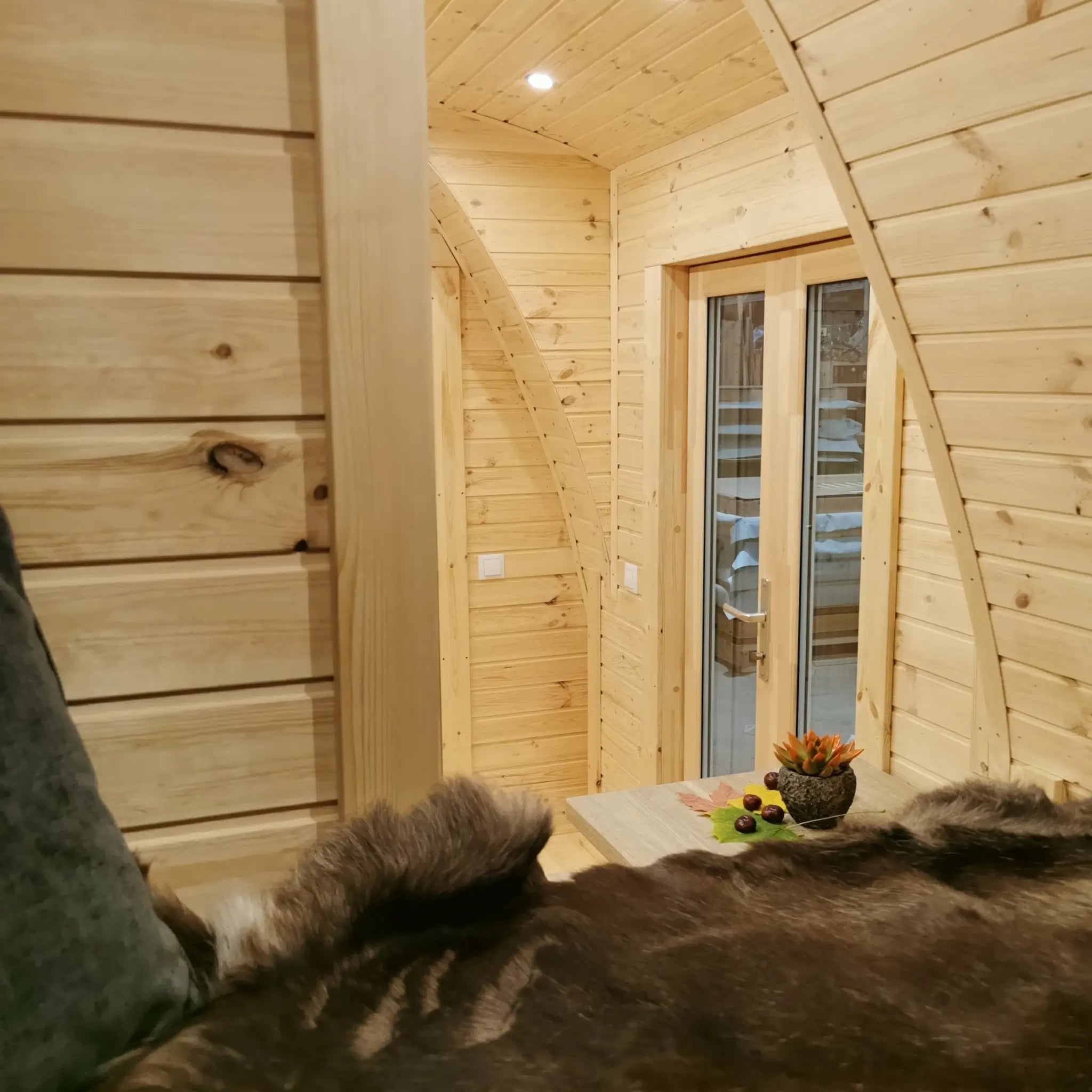 View of the entrance door from the sleeping area inside the Hearthwood Pod luxury glamping pod, showcasing curved wooden interior and warm ambient lighting.