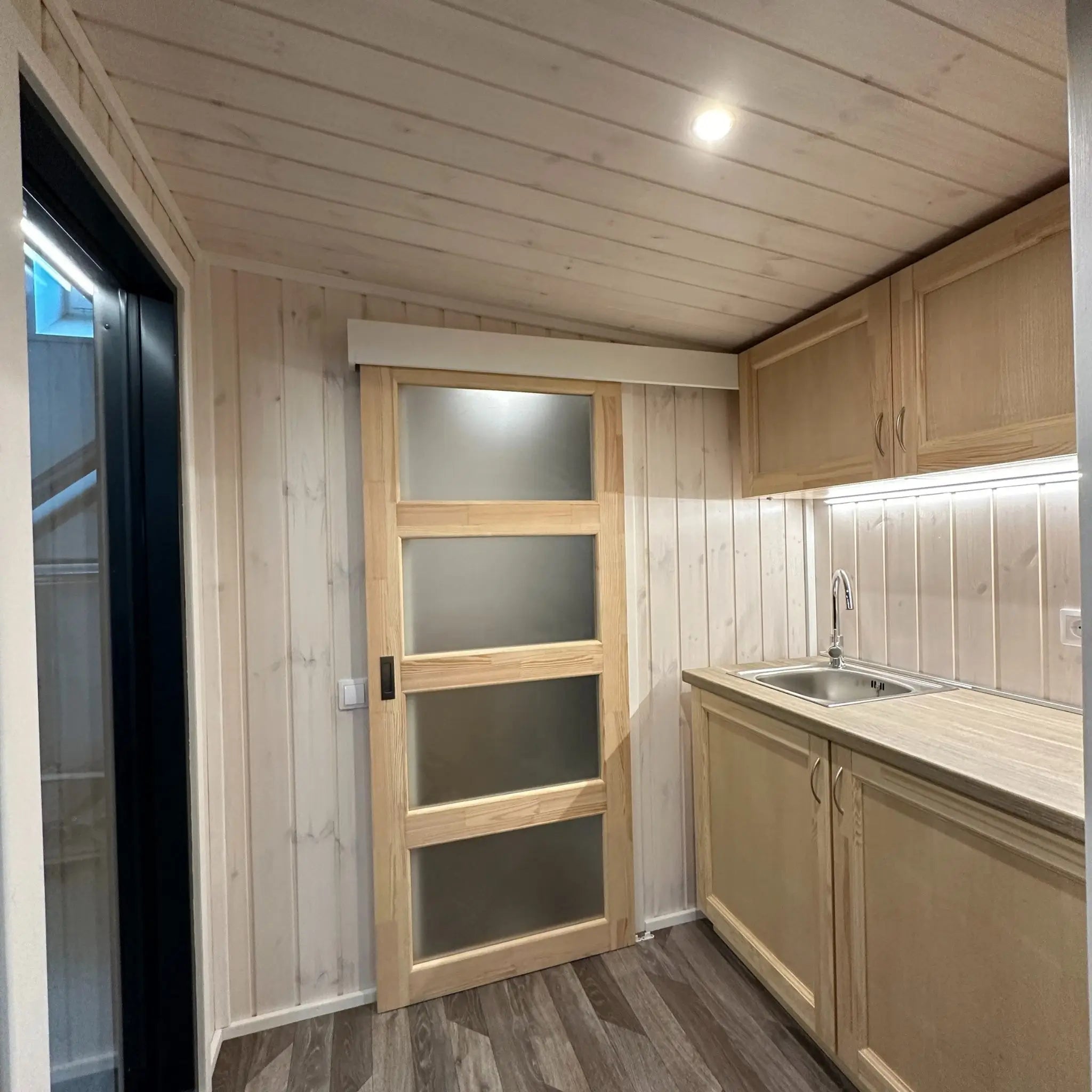 The Large GlampTainer container-style glamping unit – bright kitchenette with overhead cabinets next to closed frosted glass bathroom door