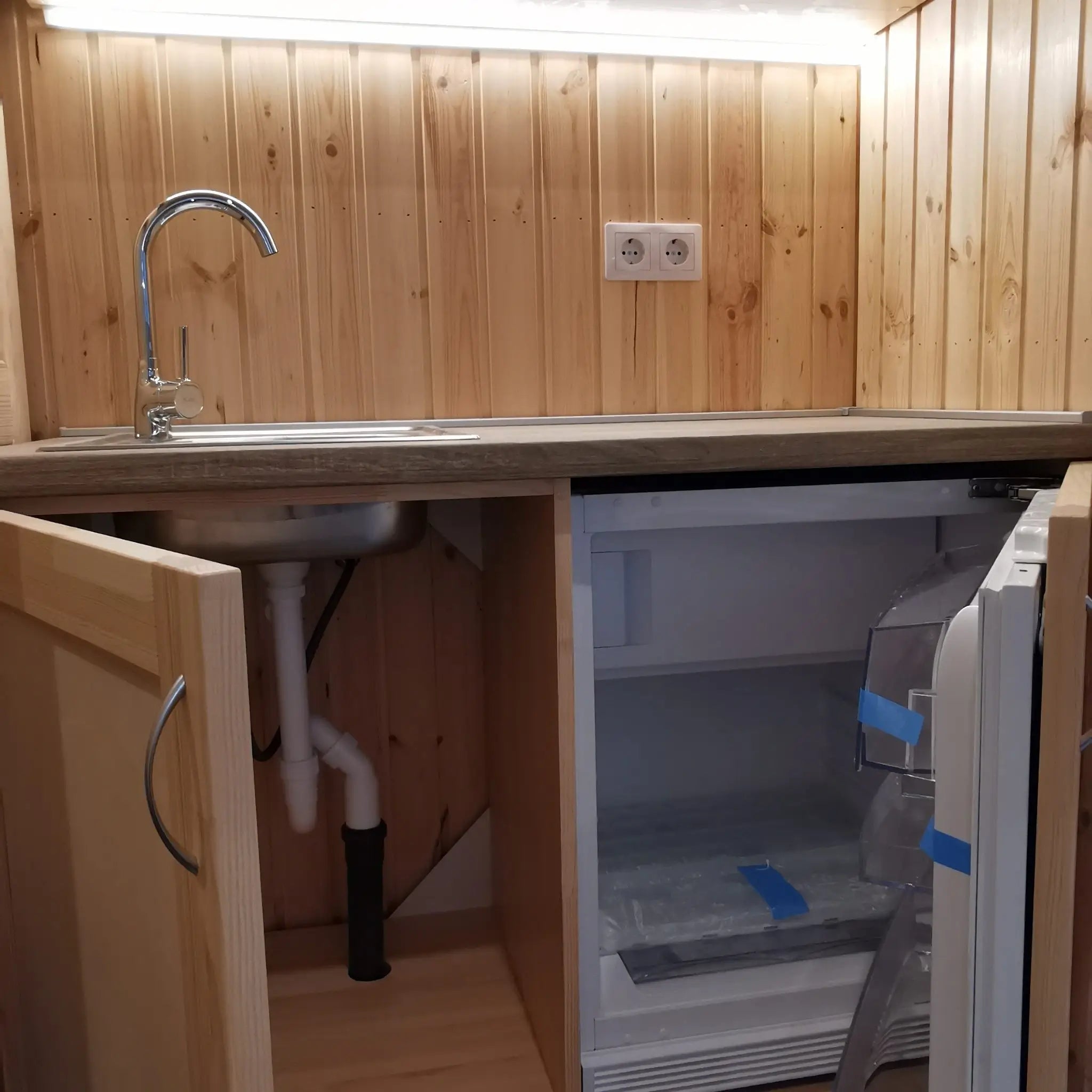 The Medium GlampTainer container-style glamping structure – compact kitchenette with open cabinets revealing under-sink storage and mini fridge