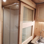 The Medium GlampTainer container-style glamping structure – compact shower cubicle with glass doors beside kitchenette sink and timber cabinetry