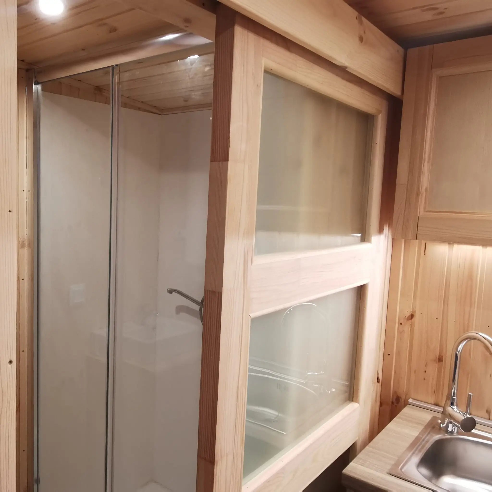 The Medium GlampTainer container-style glamping structure – compact shower cubicle with glass doors beside kitchenette sink and timber cabinetry