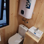 The Medium GlampTainer container-style glamping structure – space-efficient bathroom with white toilet, wall-mounted water heater and small vanity sink