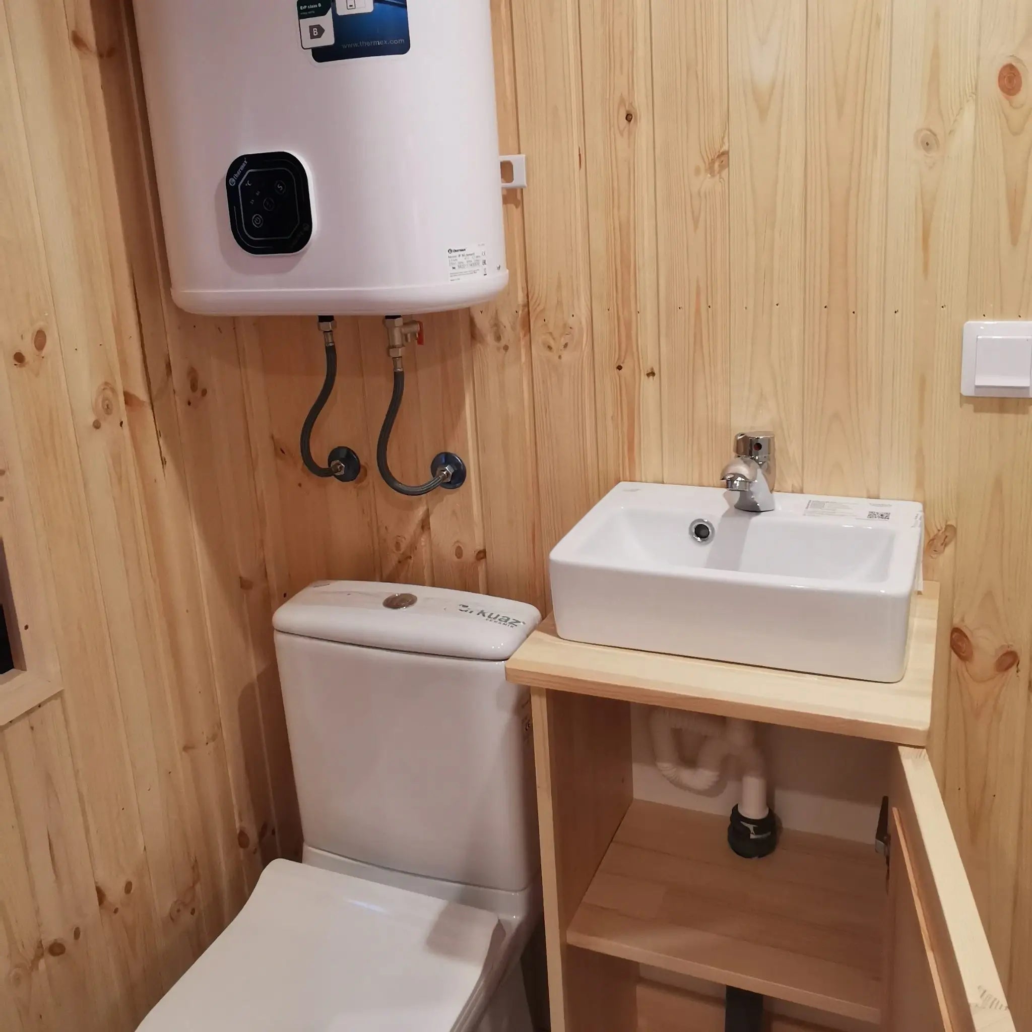 The Medium GlampTainer container-style glamping structure – compact bathroom with toilet, wall-mounted water heater and vanity cabinet open beneath basin