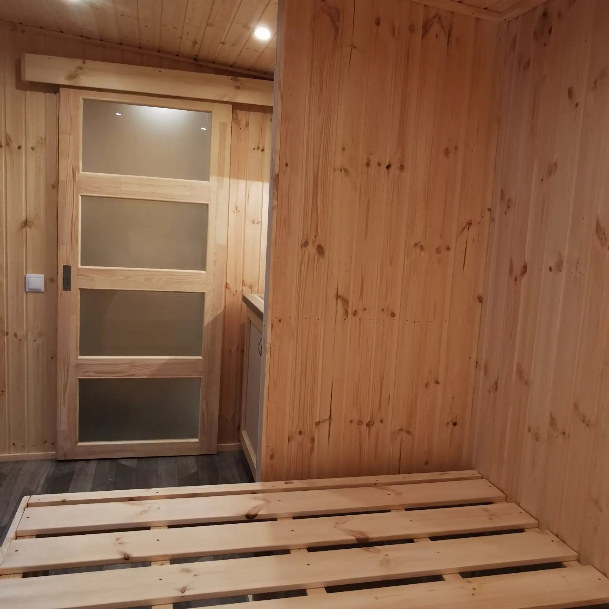 The Medium GlampTainer container-style glamping structure – wooden bed frame opposite frosted sliding door leading to bathroom beside kitchenette