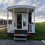 Front view of luxury shepherd’s hut with covered porch, curved roof, white decking, and modern tiny house design.