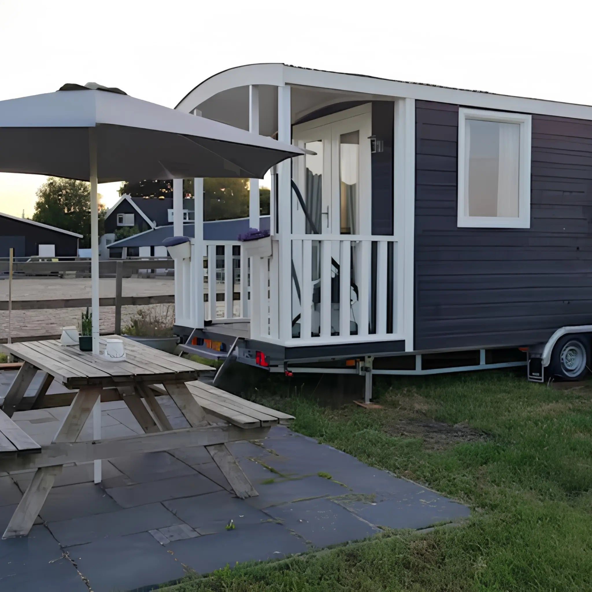 Side view of luxury shepherd’s hut beside an outdoor picnic table and parasol in a peaceful glamping setting.