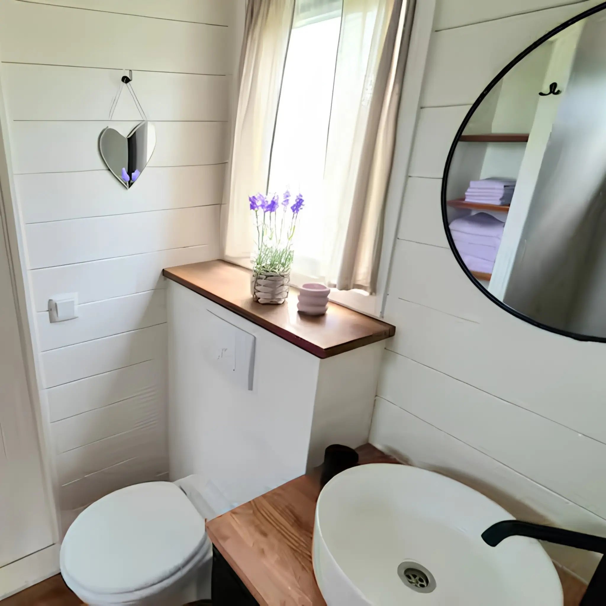 Stylish compact bathroom with round mirror, vessel sink, wooden vanity, and soft natural lighting.