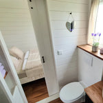 Bright bathroom interior with round mirror, wooden vanity, and doorway leading to a cosy bedroom.