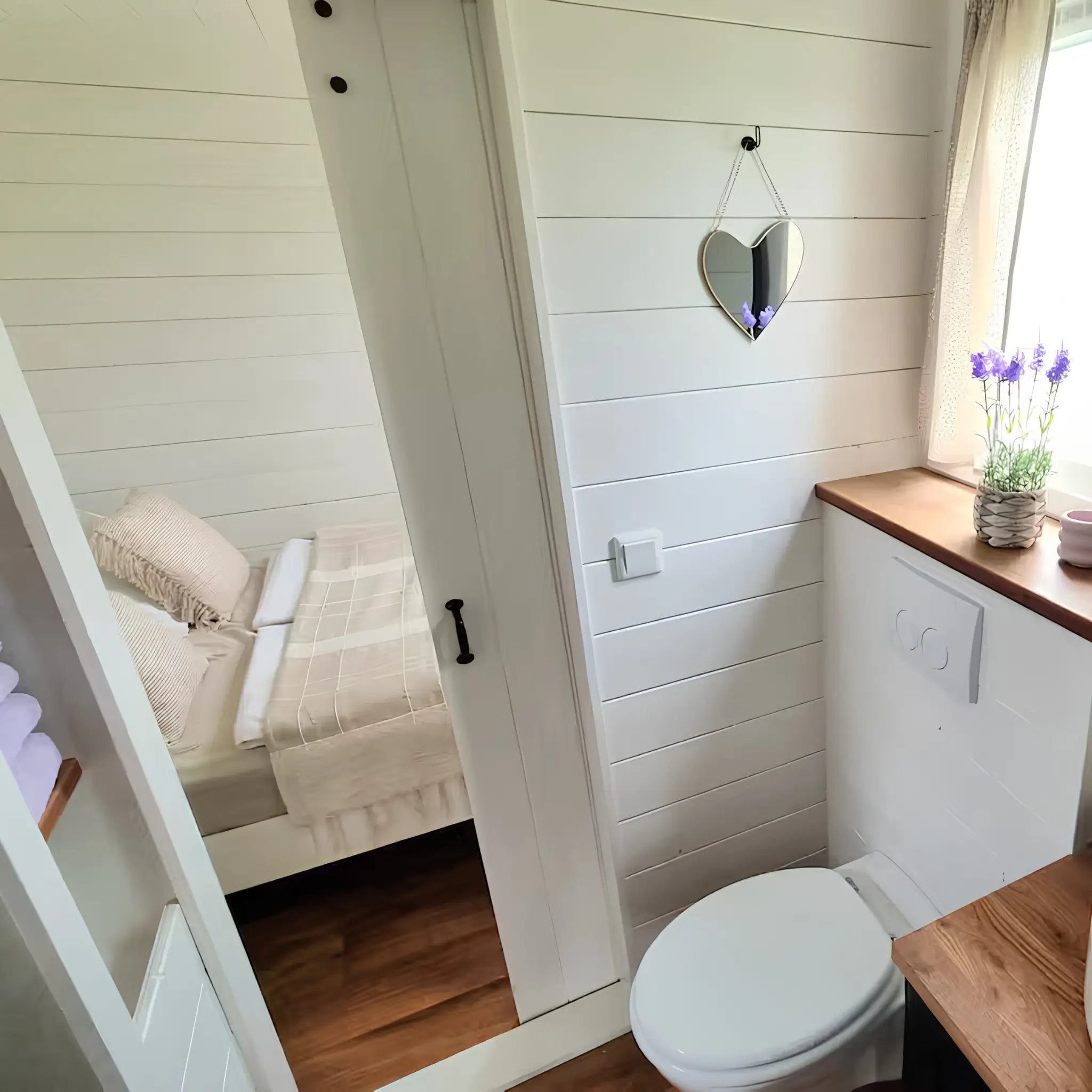 Bright bathroom interior with round mirror, wooden vanity, and doorway leading to a cosy bedroom.