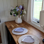 Wooden dining counter with heart-shaped plates, soft natural lighting, and floral arrangement beside a countryside window.