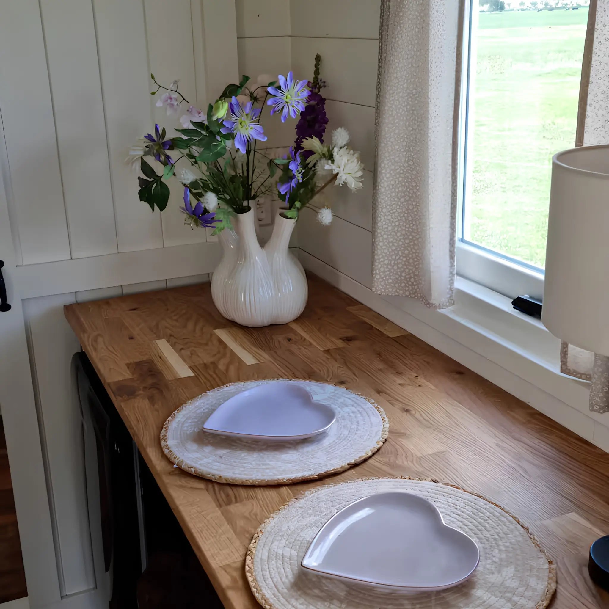 Wooden dining counter with heart-shaped plates, soft natural lighting, and floral arrangement beside a countryside window.