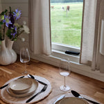 Cosy dining setup beside a large window overlooking open countryside fields, with neutral tableware, wine glasses, and floral decor.