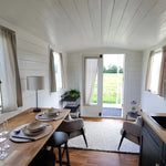 Open-plan shepherd’s hut interior with dining area, kitchenette, lounge seating, and glass doors opening to scenic countryside views.
