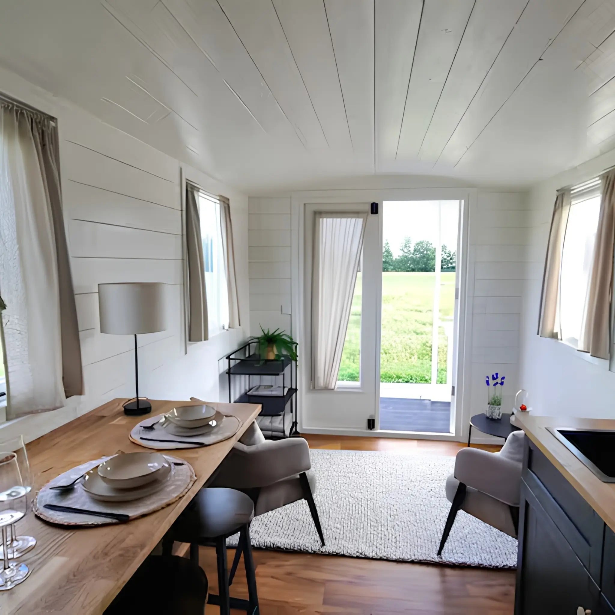 Open-plan shepherd’s hut interior with dining area, kitchenette, lounge seating, and glass doors opening to scenic countryside views.