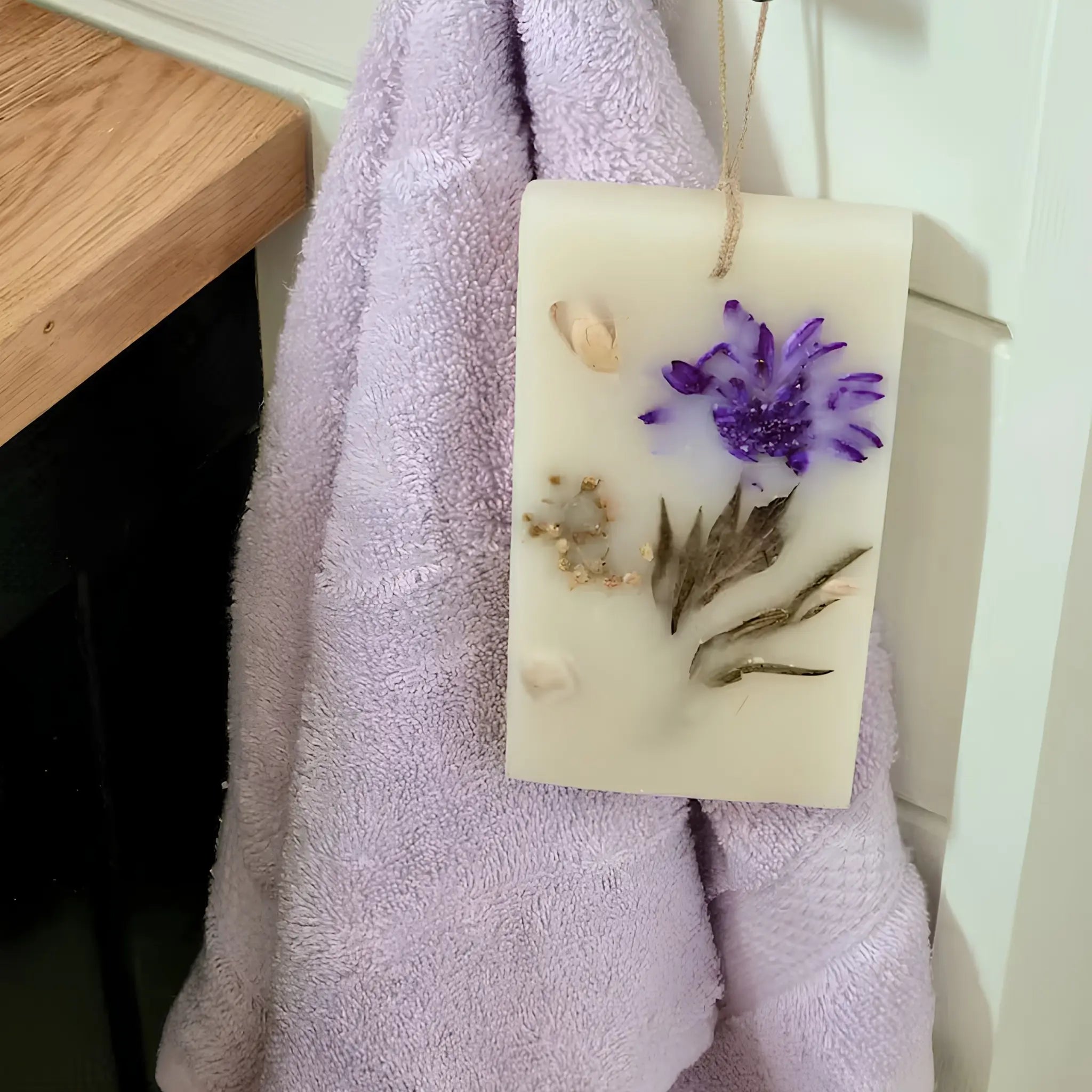 Decorative hanging soap bar with pressed purple flowers beside a soft lilac hand towel.