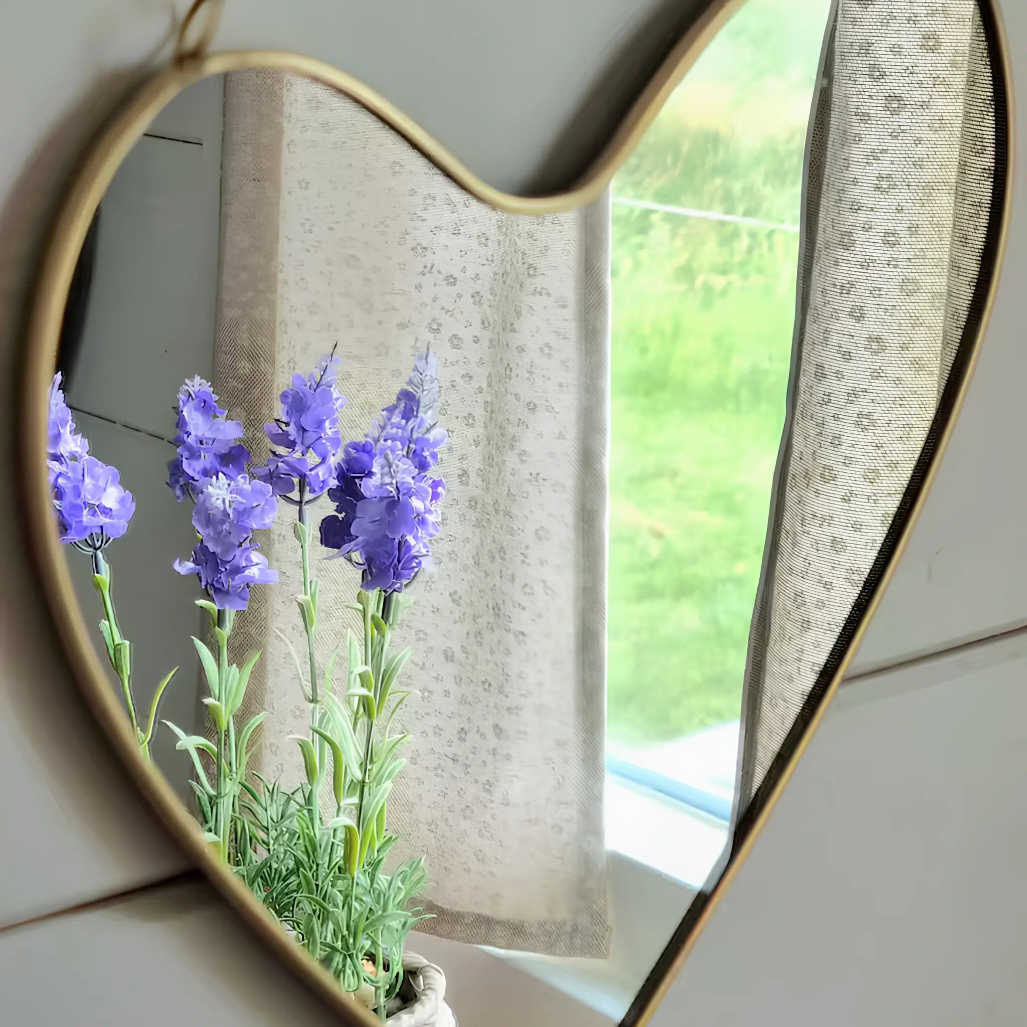 Heart-shaped wall mirror reflecting purple flowers beside a softly lit countryside window.