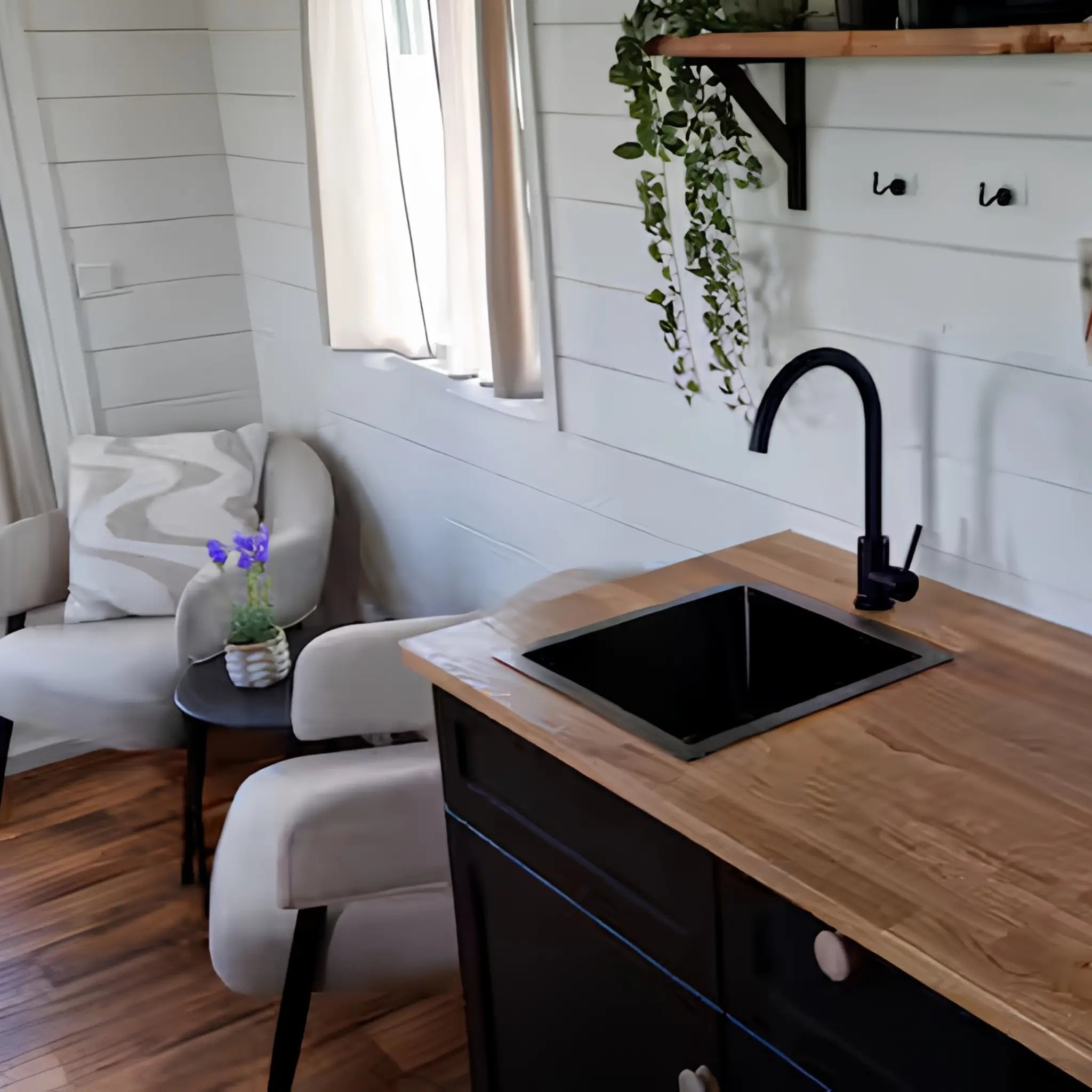 Modern kitchenette with black sink, wooden worktops, floating shelves, and cosy lounge seating nearby.