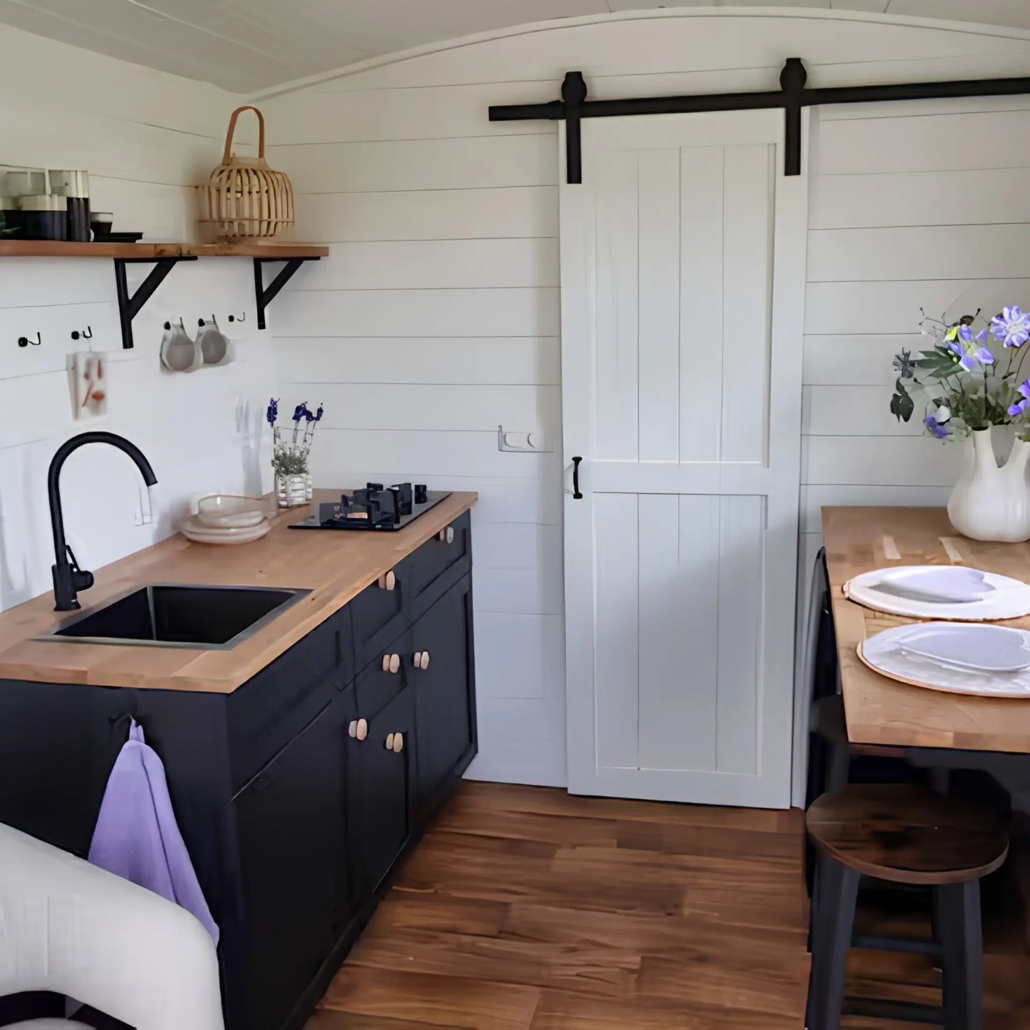 Compact kitchenette with black cabinetry, wooden countertops, floating shelves, and dining area beside a sliding barn door.