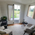 Bright lounge area with soft armchairs, textured rug, black shelving unit, and glass doors opening to countryside views.