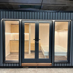 The Small GlampTainer container-style glamping structure – front view with full-glass doors revealing bright Scandi-style interior with kitchenette and bed frame.