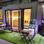 Fully furnished Small GlampTainer container-style glamping structure – showroom display with double bed, kitchenette, and outdoor bistro seating on artificial grass.