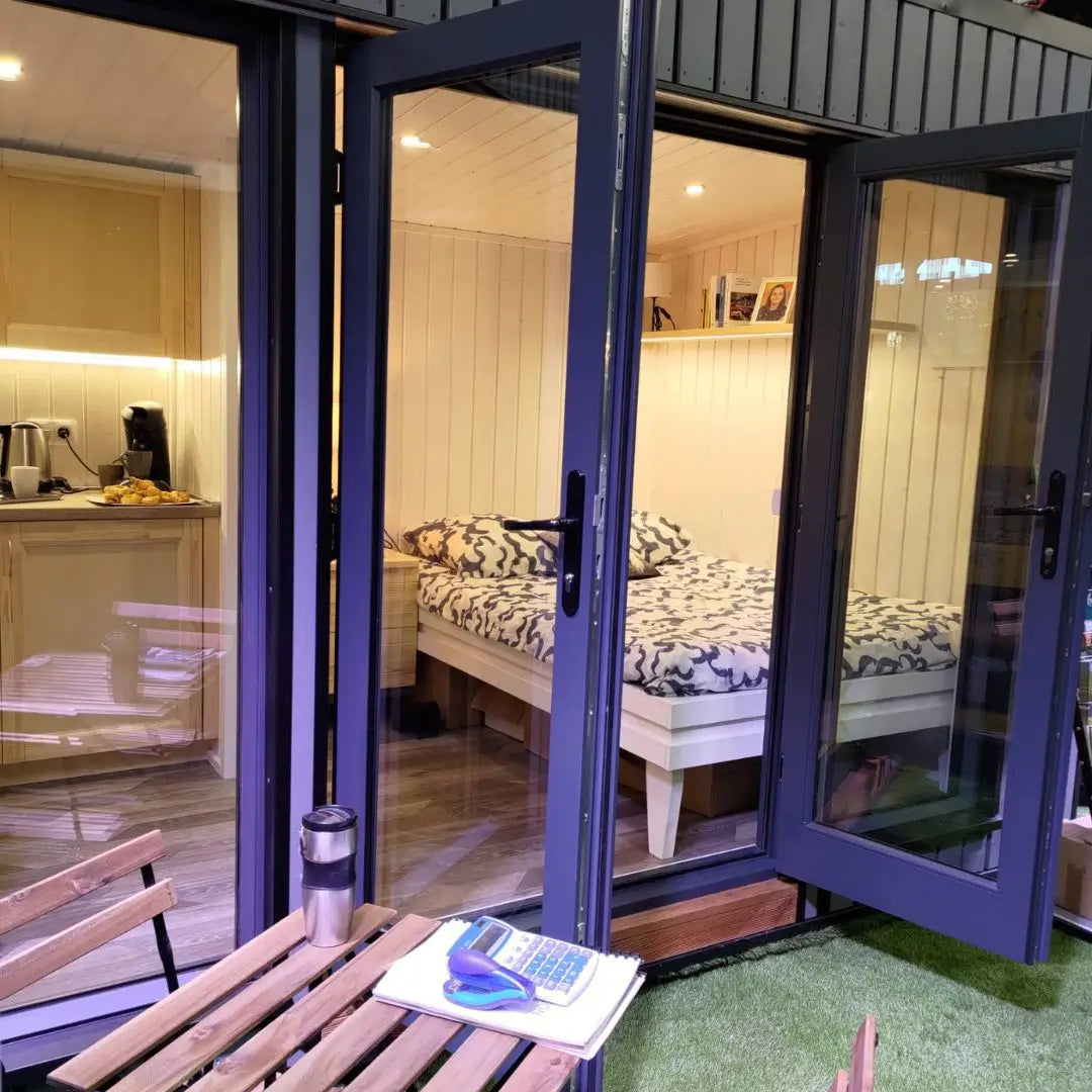 Fully furnished Small GlampTainer container-style glamping structure – cosy sleeping area with double bed, kitchenette and outdoor bistro setup on artificial grass.