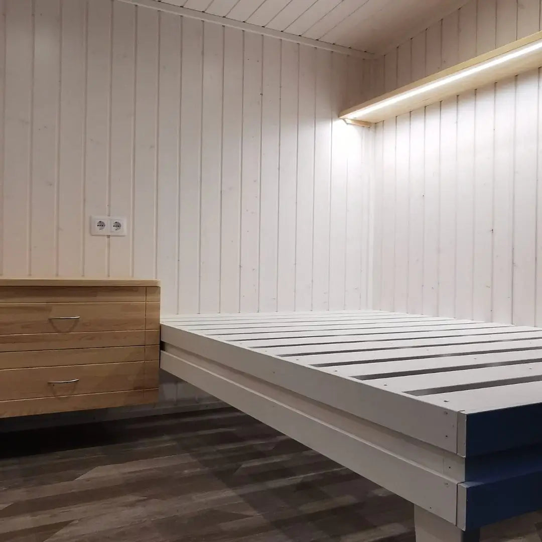 Minimalist sleeping nook in The Small GlampTainer container-style glamping structure – wooden nightstand and white bed frame against Scandi-style timber walls.