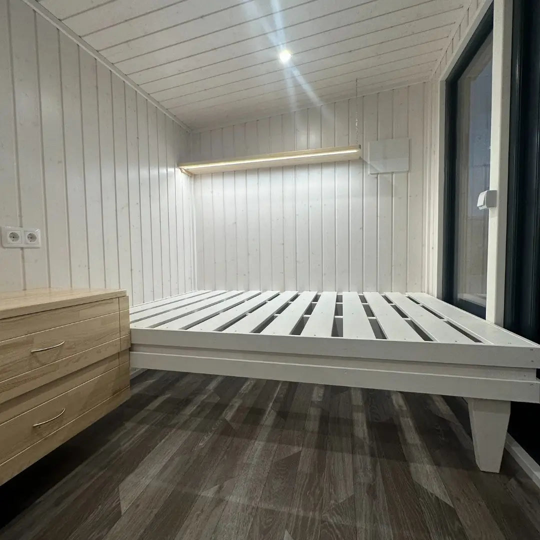 Bright sleeping area inside The Small GlampTainer container-style glamping structure – white wooden bed frame with built-in nightstand and warm strip lighting.