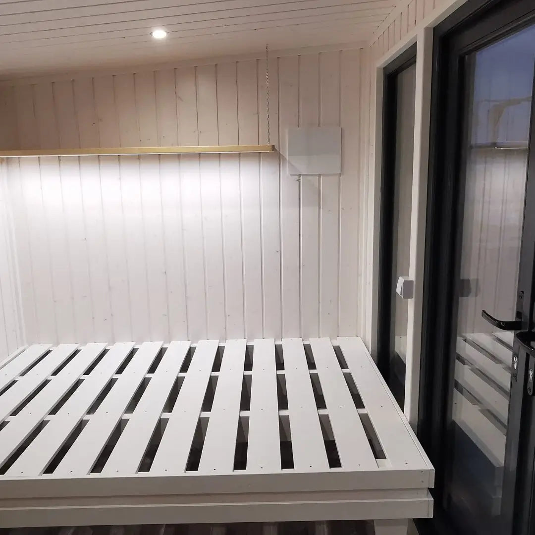 Sleeping area inside The Small GlampTainer container-style glamping structure – white wooden bed frame beside full-height glass wall and soft strip lighting.