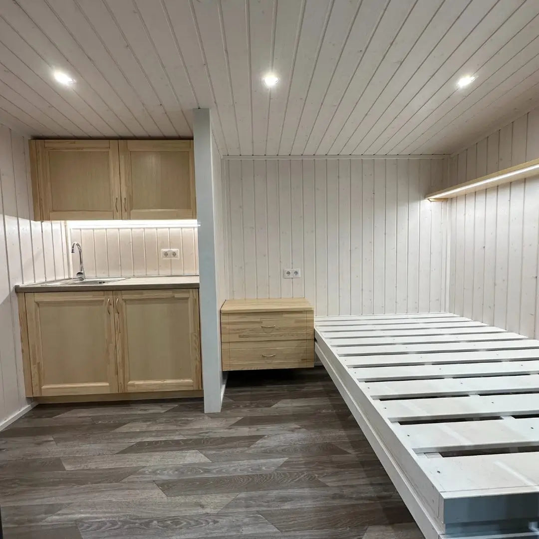 Open-plan interior in The Small GlampTainer container-style glamping structure – bright space with kitchenette, floating nightstand and wooden bed frame.
