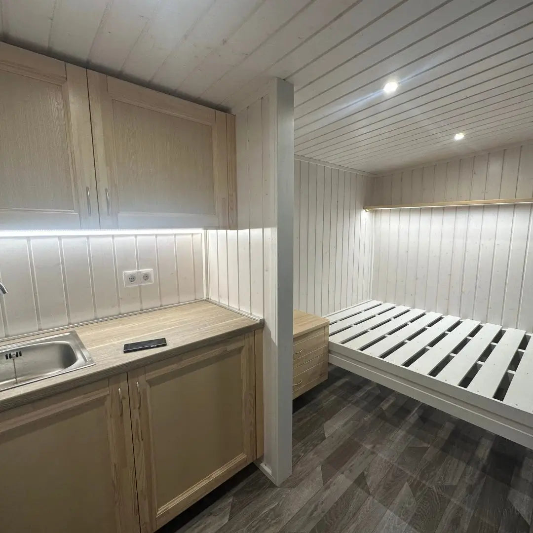 Compact interior of The Small GlampTainer container-style glamping structure – view from right side showing kitchenette, bed frame and warm downlighting.