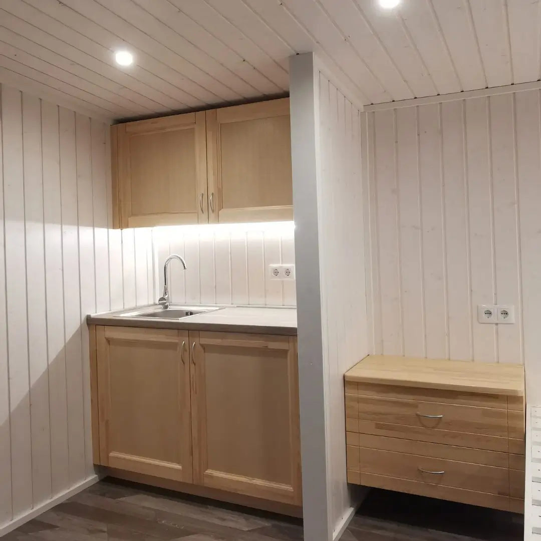 Minimalist kitchenette and nightstand inside The Small GlampTainer container-style glamping structure – light timber finishes with soft under-cabinet lighting.
