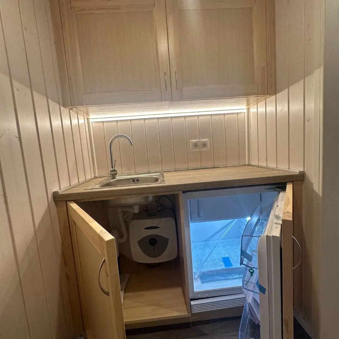 The Small GlampTainer container-style glamping structure – kitchenette with open base cupboards showing integrated mini fridge and concealed water heater unit.