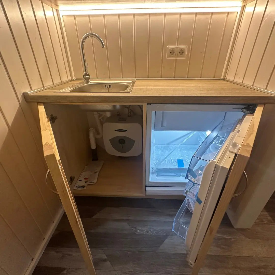 Compact kitchenette inside The Small GlampTainer container-style glamping structure – open base cabinet revealing mini fridge and built-in water heater beneath sink.