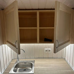 Wall-mounted cabinet in The Small GlampTainer container-style glamping structure – open double doors showing interior shelving above stainless steel sink.