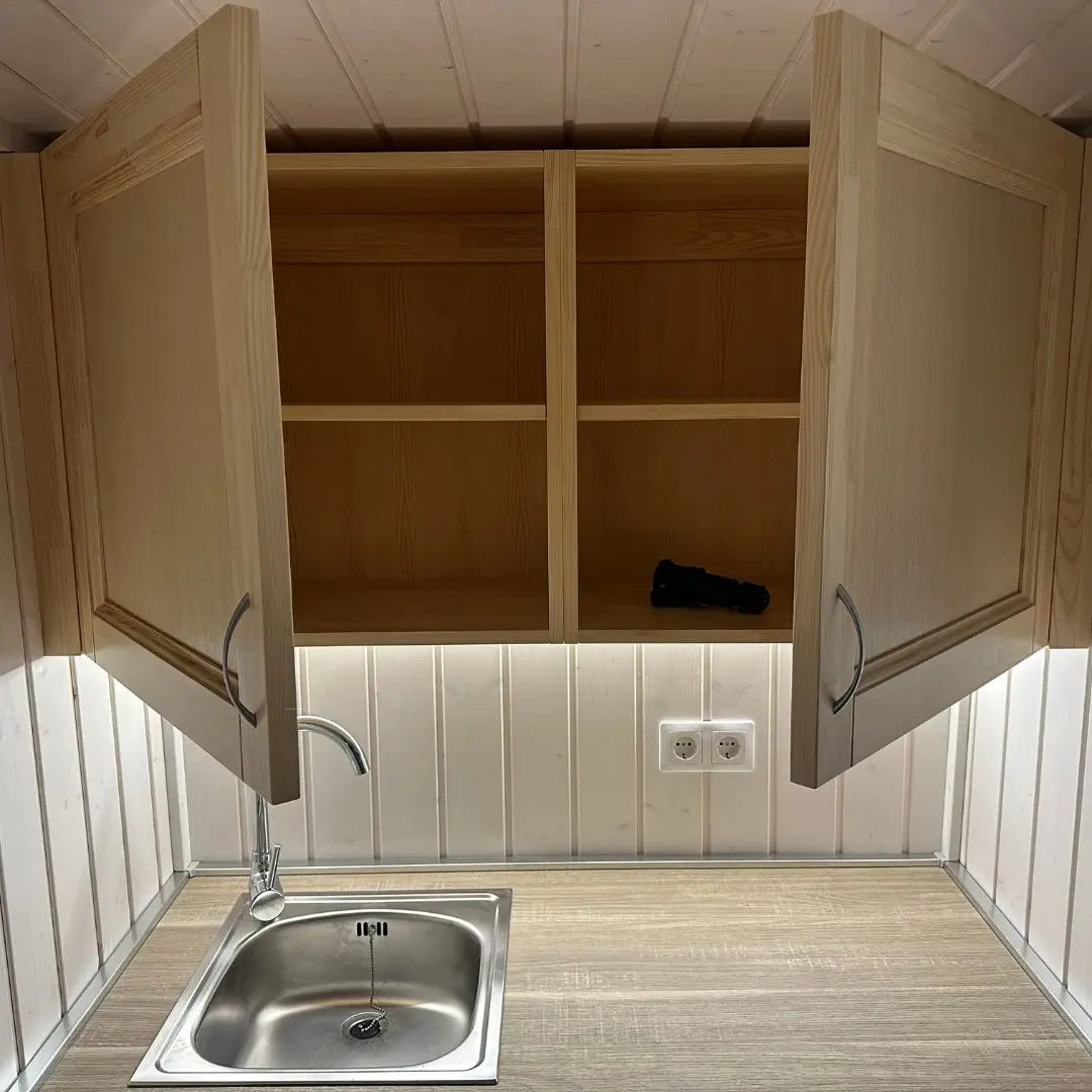 Wall-mounted cabinet in The Small GlampTainer container-style glamping structure – open double doors showing interior shelving above stainless steel sink.