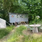 Exterior view of a cosy 2 person shepherd’s hut surrounded by lush trees, outdoor seating, and peaceful countryside greenery.