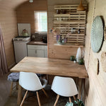 Warm wooden dining area with minimalist kitchenette, decorative shelving, and cosy seating inside a compact shepherd’s hut.