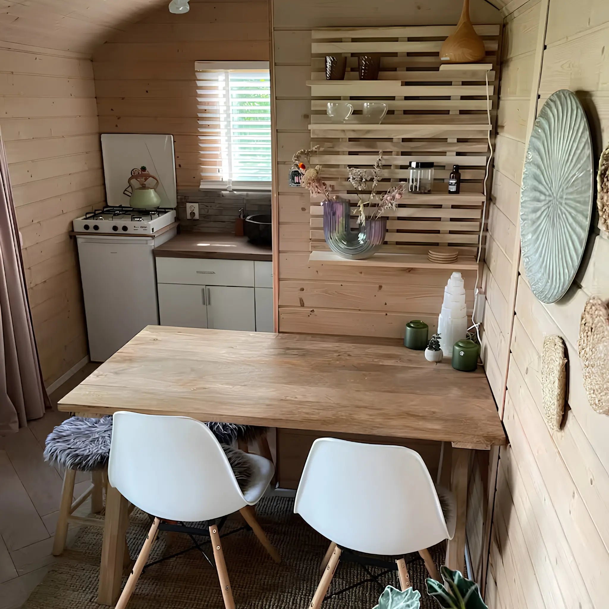Warm wooden dining area with minimalist kitchenette, decorative shelving, and cosy seating inside a compact shepherd’s hut.